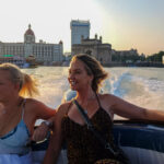 Two women smiling on a speedboat ride near the Gateway of India and Taj Mahal Palace Hotel at sunset in Mumbai.